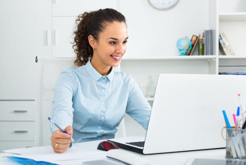 Une femme assise à un bureau, souriante devant son ordinateur portable, tient un stylo dans une main, tandis que des papiers et des fournitures de bureau sont posés sur le bureau. Des étagères et des livres sont visibles en arrière-plan.