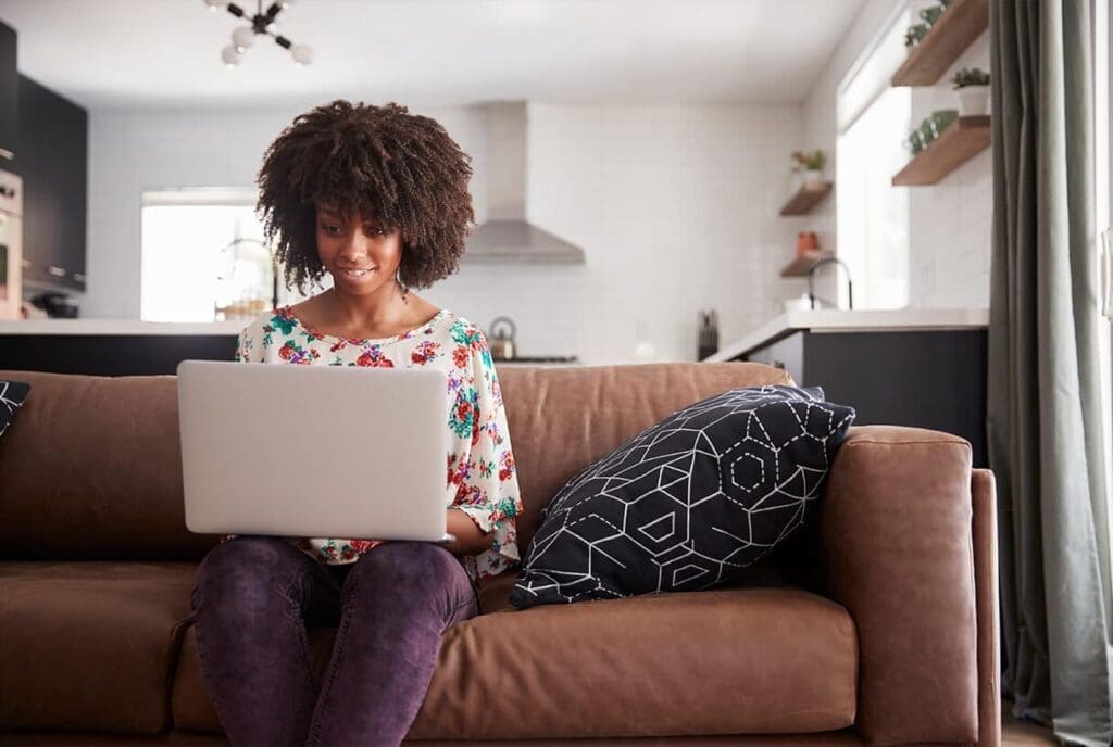 Une femme est assise sur un canapé marron dans un salon moderne, souriante, son ordinateur portable posé sur ses genoux. Un coussin à motifs est posé à côté d'elle et une cuisine est visible en arrière-plan.