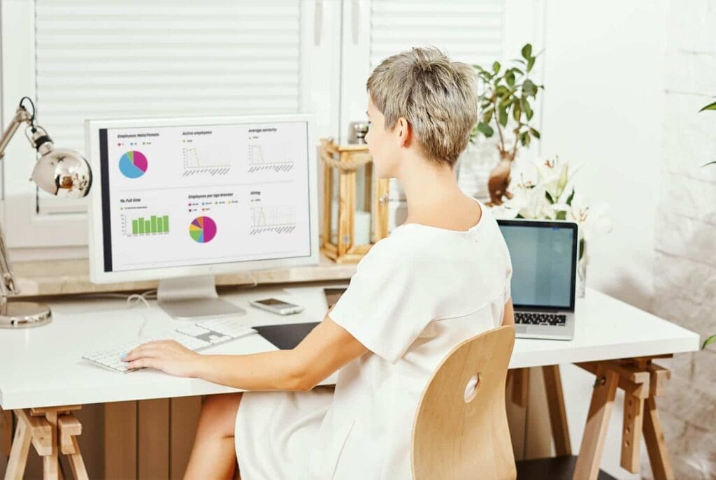 Une femme aux cheveux courts est assise à un bureau, regardant des tableaux et des graphiques colorés sur un écran d'ordinateur. Un ordinateur portable, un carnet, un téléphone et une lampe de bureau sont également posés sur le bureau blanc, dans un environnement de bureau lumineux.
