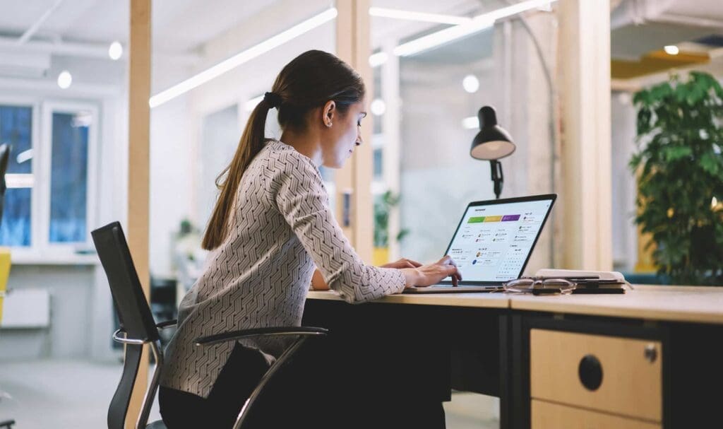 Une femme coiffée d'une queue de cheval est assise à un bureau dans un bureau moderne, travaillant sur un ordinateur portable. Une lampe de bureau, un téléphone et un carnet sont à proximité, tandis que de grandes fenêtres et des plantes sont visibles en arrière-plan.