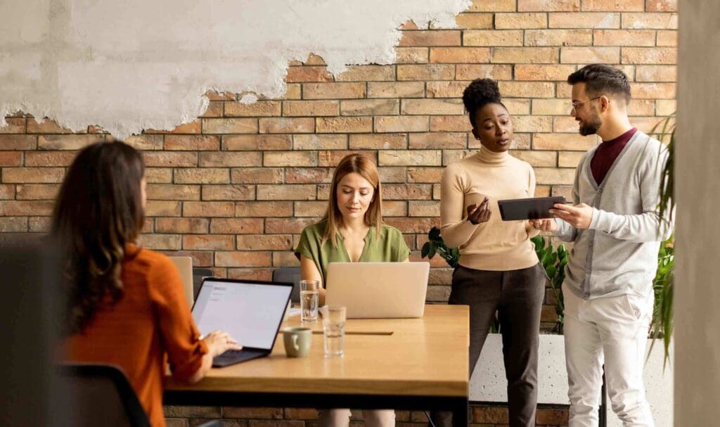 Quatre personnes dans un bureau moderne : deux assises à une table avec des ordinateurs portables, et deux debout discutant sur une tablette numérique devant un mur de briques. Des verres d'eau et des cahiers sont posés sur la table.