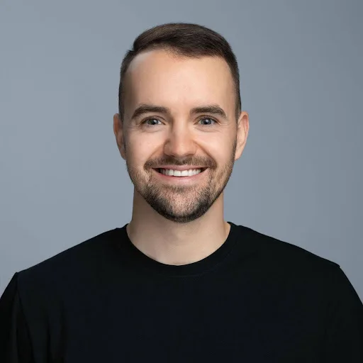 A smiling man with short brown hair and a beard, wearing a black crew-neck shirt, poses against a plain gray background.