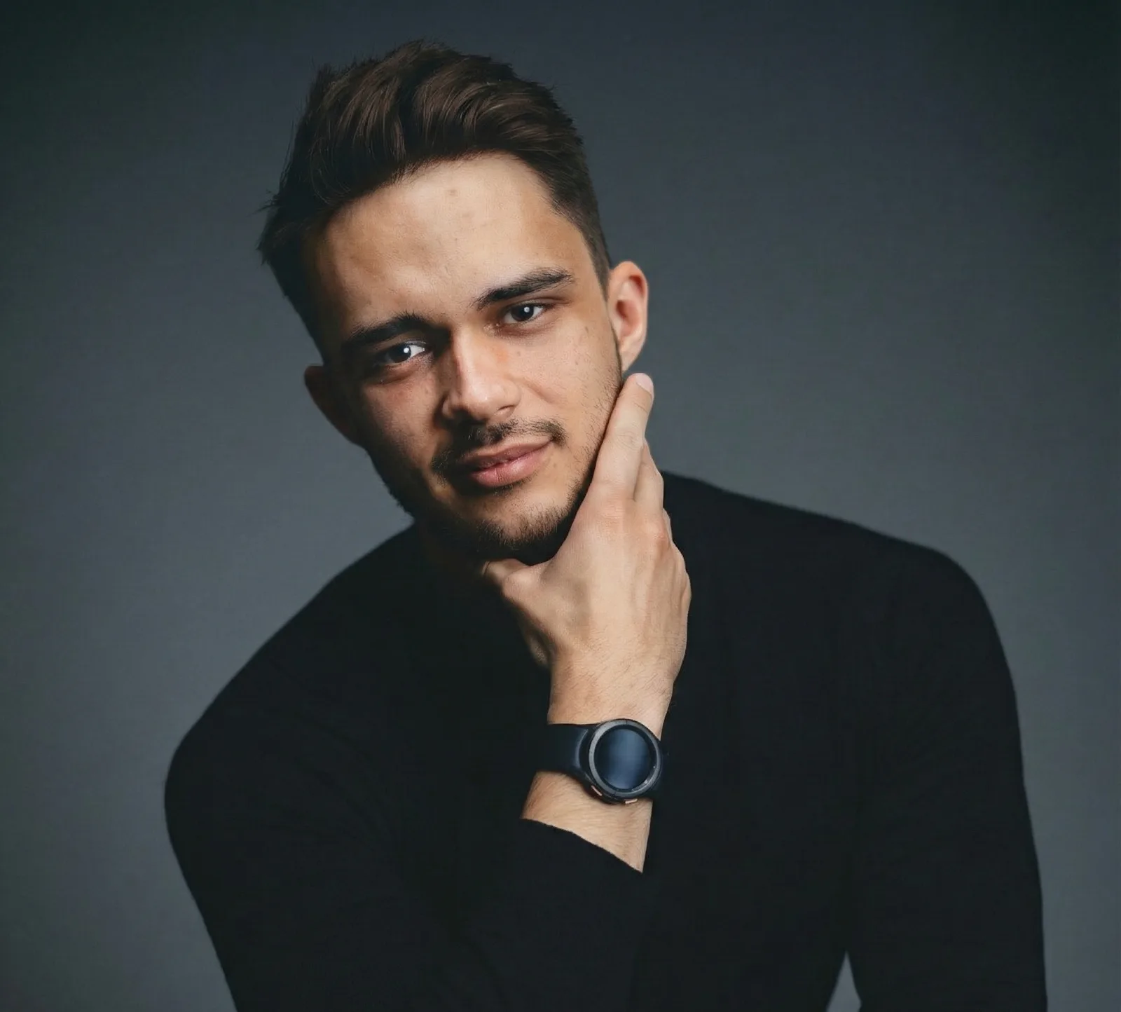 A young man with short brown hair and facial hair wears a black long-sleeve shirt and a black wristwatch, resting his chin on his hand and looking confidently at the camera against a dark background.