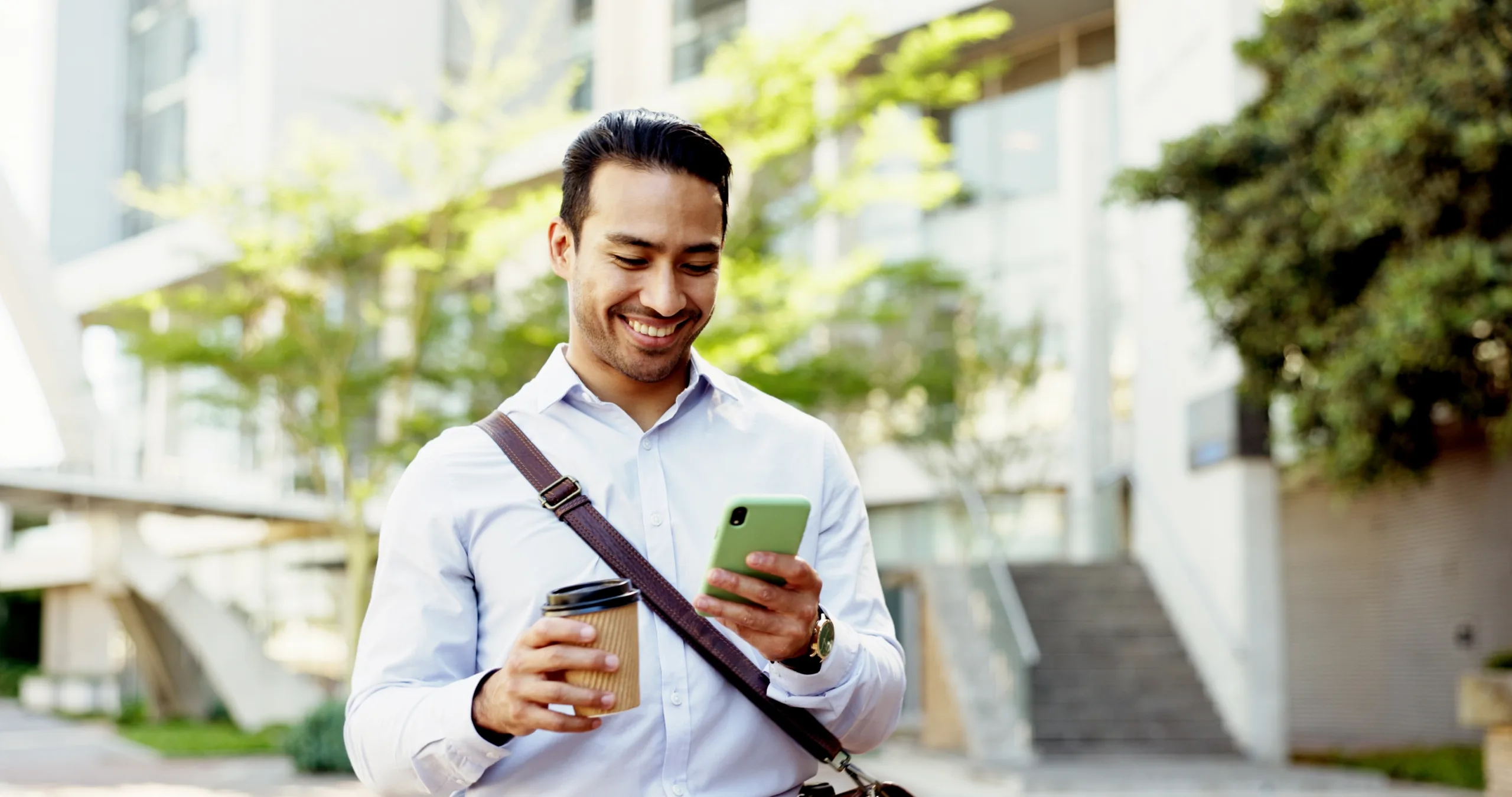 Smiling man outdoors in business attire holds a coffee cup in one hand and looks at his smartphone in the other, with a satchel over his shoulder and greenery in the background.