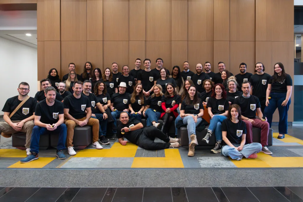 A large group of people wearing matching black T-shirts with a crest logo pose together indoors, some seated, some standing, and a few sitting on the floor, smiling in front of a wooden wall panel.